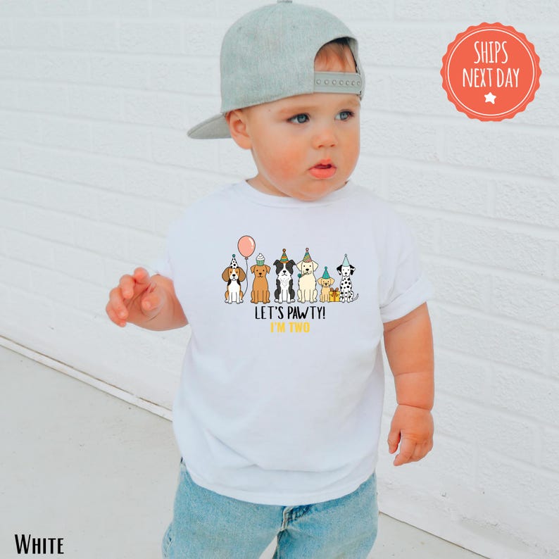 A young child wearing a white t-shirt with a graphic of dogs and a birthday party theme, standing in front of a white brick wall.