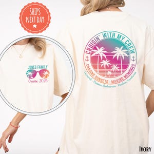 A person wearing a white t-shirt with a colorful graphic design featuring a beach scene with palm trees and a sunset. The text on the t-shirt reads "CRUISIN' WITH MY CREW" and "JONES FAMILY Cruises 2026".