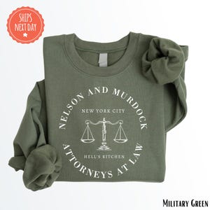 May include: Military green sweatshirt with a white graphic featuring the text "NELSON AND MURDOCK ATTORNEYS AT LAW" around a scale of justice. The text "NEW YORK CITY" and "HELL'S KITCHEN" are also included.