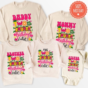 May include: A collection of beige sweatshirts and a baby bodysuit featuring colorful fruit-themed graphics. Each garment displays the text "Twotti Frutti Birthday Cutie" alongside illustrations of oranges, watermelons, pineapples, and pink flamingos.