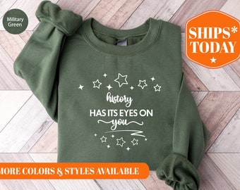 May include: A green sweatshirt with the text "history has its eyes on you" in white lettering surrounded by white stars. The sweatshirt is on a wood background.