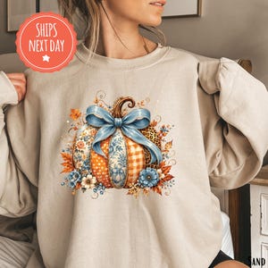 May include: A beige crewneck sweatshirt featuring a central graphic of a pumpkin decorated with various patterns including floral, gingham, polka dot, and leopard print. A large light blue bow sits atop the pumpkin, surrounded by autumn leaves.