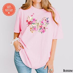 A woman wearing a pink t-shirt with a floral design and a pink butterfly on it, standing against a white background.