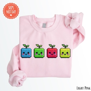 May include: Light pink sweatshirt featuring four pixelated, smiling, square-shaped characters in blue, green, red, and yellow. Each character has a small green leaf on top. The sweatshirt is a solid light pink color.