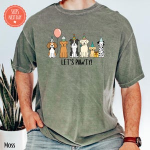 A person wearing a gray t-shirt with a graphic design of six dogs wearing party hats and the text "Let's pawty!"