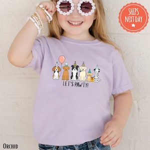 A young girl wearing a purple t-shirt with a graphic of cartoon dogs and the text "Let's pawty!"