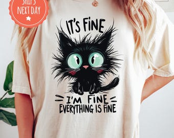 May include: An ivory t-shirt featuring a graphic of a fluffy black cat with large teal eyes. The text above the cat reads IT'S FINE and the text below reads I'M FINE EVERYTHING IS FINE in black lettering. The cat has messy fur.