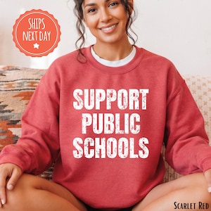 May include: A scarlet red crewneck sweatshirt featuring the white distressed text SUPPORT PUBLIC SCHOOLS centered on the chest. The garment has long sleeves, a ribbed neckline, and a classic fit, providing a simple and bold graphic design.