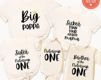May include: A collection of five off-white shirts and a bodysuit featuring black text and small gold crowns. The garments display the phrases Big Poppa, Sicker Than Your Average Mama, Sister of the Notorious ONE, Notorious ONE, and Brother of the Notorious ONE.