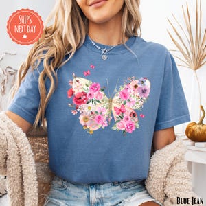 A woman wearing a blue t-shirt with a colorful butterfly design and floral patterns, sitting in a room with a potted plant and a stuffed animal.