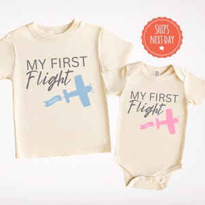 May include: Two cream colored shirts with the text "My First Flight" and a small airplane graphic. The shirt on the left has a blue airplane and the text "Andi" on a banner. The shirt on the right has a pink airplane and the text "Abi" on a banner.
