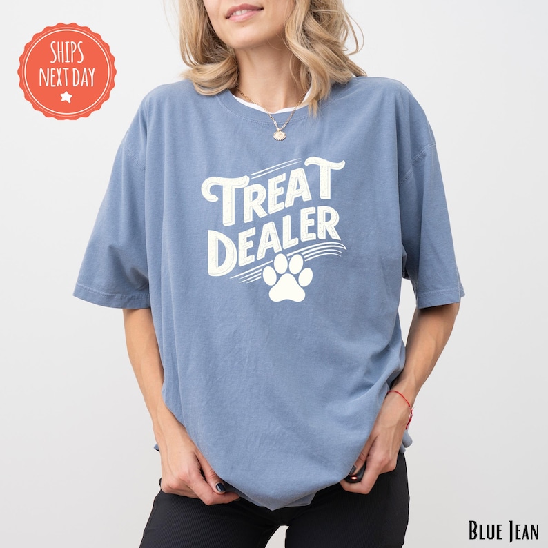 K&ouml;nnte beinhalten: Blaues T-Shirt mit wei&szlig;em Schriftzug "TREAT DEALER" und wei&szlig;em Pfotenabdruck. Das Shirt hat eine lockere Passform und einen Rundhalsausschnitt. Der Text "BLUE JEAN" ist unten rechts aufgedruckt.