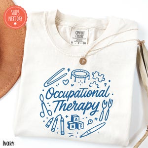 a white t-shirt with a blue design that says "Occupational Therapy" and includes various objects such as scissors, a pencil, and a stapler.