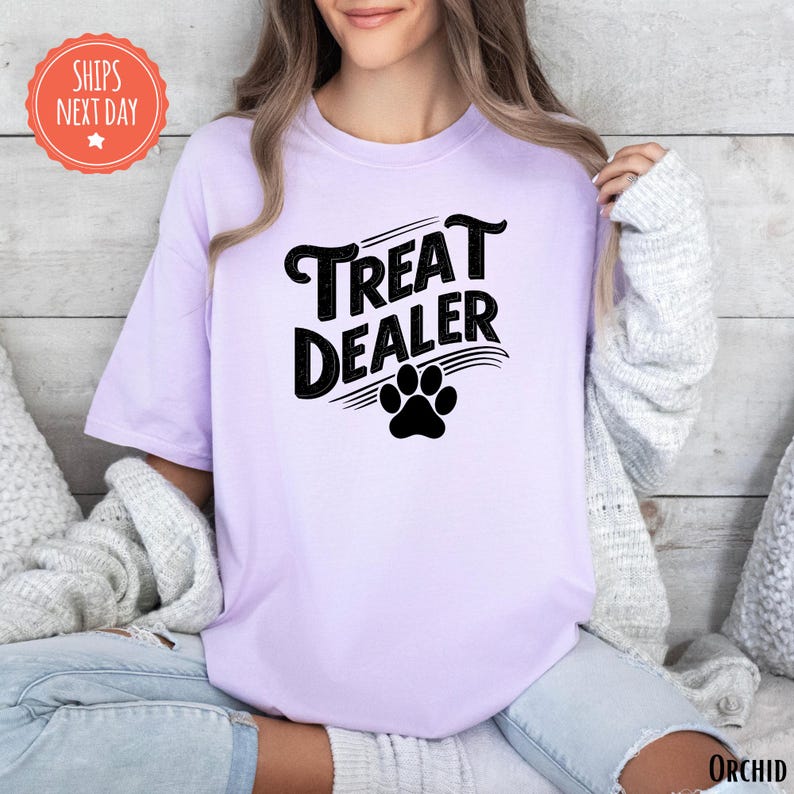 K&ouml;nnte beinhalten: Helllila T-Shirt mit dem schwarzen Schriftzug "TREAT DEALER" und einem Pfotenabdruck. Das Shirt hat kurze &Auml;rmel und eine lockere Passform. Ein orangefarbener Aufkleber mit "SHIPS NEXT DAY" ist ebenfalls zu sehen.