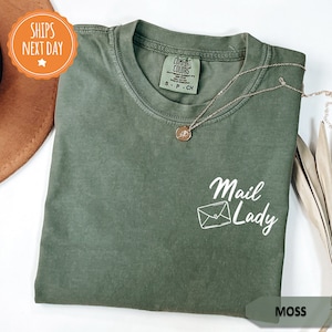 May include: A moss green t-shirt with a white graphic of an envelope and the text "Mail Lady".