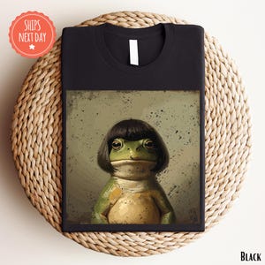 May include: Black t-shirt featuring a whimsical illustration of a frog with a black bob haircut. The frog is depicted in shades of green and tan, set against a muted background. The shirt is displayed on a woven surface, with a "Ships Next Day" sticker.