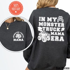 May include: Dark heather gray sweatshirt with white text and graphics. The front features a small design that says "Monster Truck Mama." The back of the sweatshirt says "In My Monster Truck Mama Era" with a monster truck graphic.