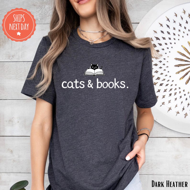 A woman wearing a gray t-shirt with the text "cats & books" printed on it, standing in front of a wooden wall with a circular design.