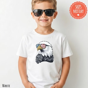 May include: White t-shirt featuring a graphic of an eagle wearing sunglasses with an American flag design. The eagle has white and black feathers. The text "WHITE" is at the bottom left of the shirt.