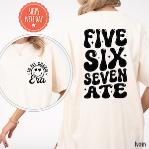 May include: Ivory-colored t-shirt with the text "FIVE SIX SEVEN ATE" in black on the back. The front of the shirt has a circular graphic with the words "IN MY CHEER Era" and a smiley face. The shirt is a casual style.