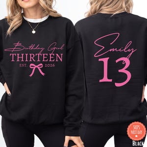 May include: A black crewneck sweatshirt featuring pink text and graphics. The front displays the words Birthday Girl, THIRTEEN, EST. 2026, and a bow icon. The back shows the name Emily in script above the large number 13 in a bold pink font.