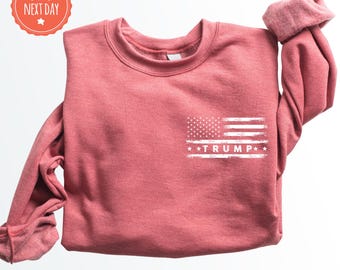 May include: A red sweatshirt with a white distressed American flag and the text "TRUMP" printed on the front. The sweatshirt is a crew neck style.
