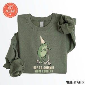 May include: Military green sweatshirt featuring a cartoon frog wearing a dunce cap and riding a scooter. The frog has a serious expression and the text "OFF TO COMMIT MOM FOOLERY" is printed below the frog.