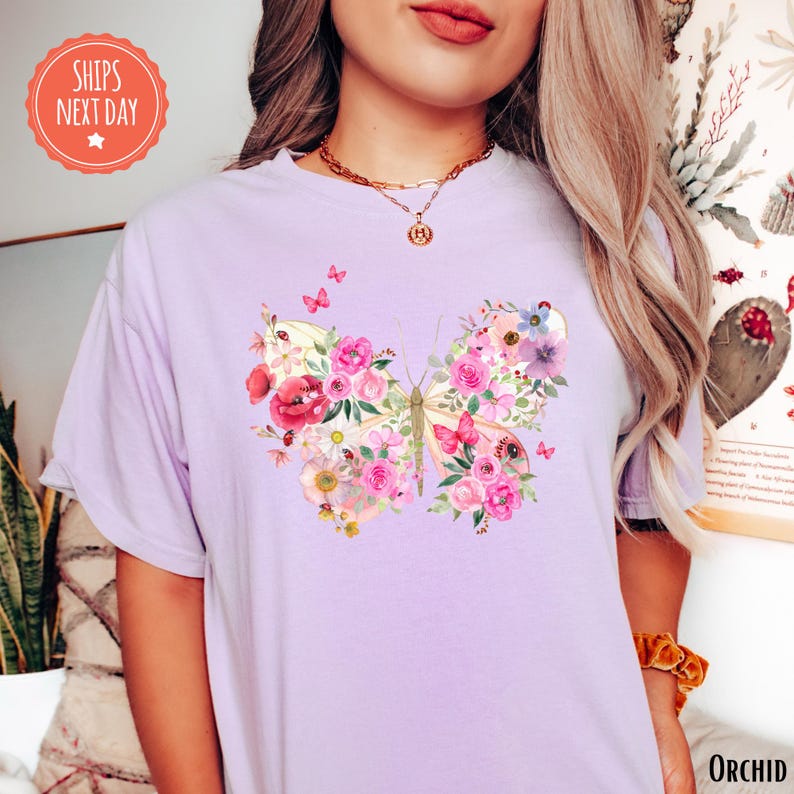 A woman wearing a purple t-shirt with a floral butterfly design, standing in front of a plant and a wall with a sign.