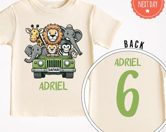 Può includere: Una maglietta beige con una jeep verde che trasporta un gruppo di animali della savana. La jeep ha una targa che dice "SAFARI". La maglietta ha il nome "ADRIEL" stampato sul davanti e il numero "6" sul retro.