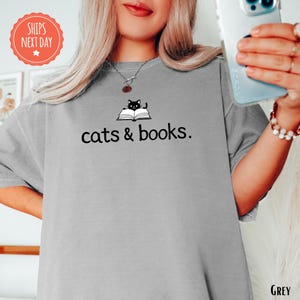 a woman holding a cell phone and wearing a gray shirt with cats and books on