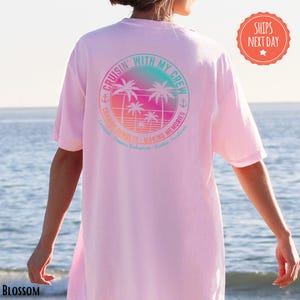 A person wearing a pink t-shirt with a graphic design of palm trees and a sunset, standing in front of a body of water.