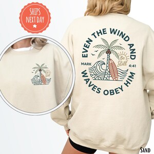 May include: Sand-colored sweatshirt with a palm tree, surfboard, and wave graphic. The back of the sweatshirt features the text "Even the wind and waves obey Him" in a circular design. The front has a smaller version of the design.