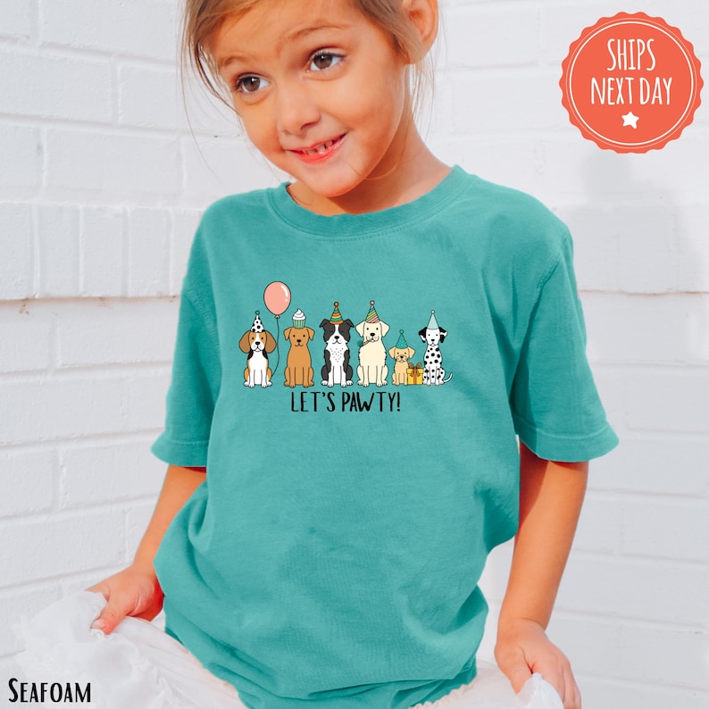 A young girl wearing a teal shirt with a graphic of cartoon dogs and the text "Let's Pawsy!"