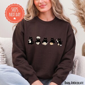 May include: A dark chocolate brown crewneck sweatshirt featuring a graphic print of five black cats engaged in baking activities, including mixing, rolling dough, and decorating a cupcake, arranged in a horizontal line across the chest.