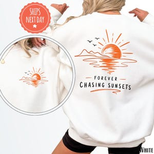 May include: White sweatshirt with an orange sunset graphic on the back. The design includes a sun, mountains, water, and the text "FOREVER CHASING SUNSETS." A smaller version of the graphic is on the front. The sweatshirt is a casual style.