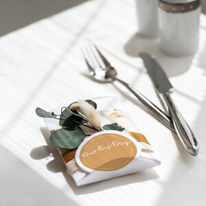 May include: A white favour box with a gold ribbon and a sticker reading "Our Big Day". The box is decorated with green leaves and dried flowers. A silver fork and knife are placed nearby.