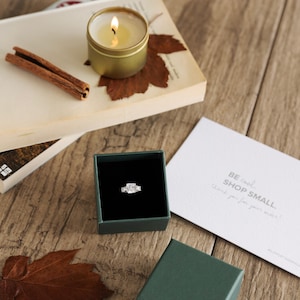 May include: A diamond ring in a dark green box, with a lit gold candle, cinnamon sticks, and a card that says "BE cool SHOP SMALL". The items are on a wooden surface.
