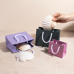 May include: Three coloured gift bags, in purple, dark green, and pink, with white rope handles. Seashells are placed inside and around the bags. A hand is placing a seashell into the dark green bag.