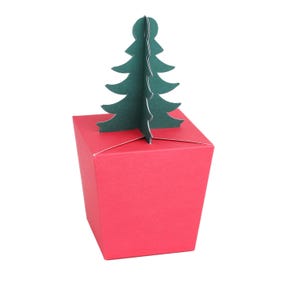 May include: A red gift box with a green Christmas tree decoration. The box is square with a folded lid. The tree is a simple silhouette with layered branches. The background is white.
