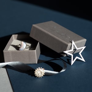 May include: A silver ring featuring a pearlescent moonstone shaped like a crescent moon. The ring is presented in a grey box with the words "The Moon Room" printed on the lid. A silver star and a silver sun charm are also visible in the image.