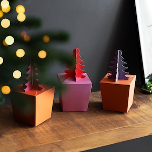 May include: Three colourful, geometric gift boxes with paper Christmas tree toppers. The boxes are orange, pink, and orange, with the trees in red and purple. The boxes are arranged on a wooden surface.