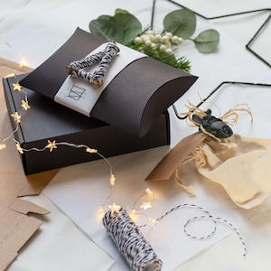 May include: A dark grey pillow box with a white band and a black and white twine bow. A dark grey box is beneath the pillow box. A string of star-shaped fairy lights and a spool of twine are also visible. The scene is set on a white surface.