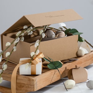 May include: A brown cardboard gift box with a lid that says "lovely the little hamper company" is open and filled with a white bowl of speckled eggs. The box is sitting on a wooden surface with a white gift box wrapped in gold ribbon, a sprig of greenery, and a brown paper apple-shaped box.