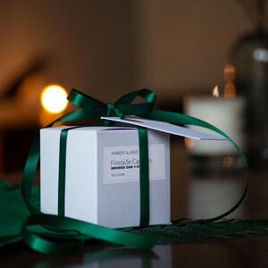 May include: A white square gift box tied with dark green satin ribbon and a bow. The box has a label that reads "Amber & Jade Fireside Candles Smoked Oak + C". A small white tag is attached to the bow. A lit candle is in the background.