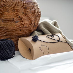 May include: A brown cardboard gift box with a black yarn ball and paw print design. A black yarn is attached to the ball and box. A woven basket and a beige blanket are in the background. A black yarn ball is in the foreground.
