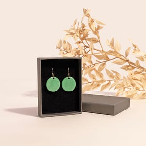 May include: A pair of green circle earrings in a black box with a grey lid. The earrings are on a black background and have gold hooks.