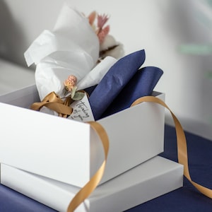 May include: White gift box with a gold ribbon and a tag that says "Thinking of you". The box is filled with blue tissue paper and dried flowers.