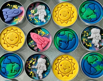 以下が含まれることがあります： 宇宙をテーマにしたデザインのカラフルな粘土が入った小さな丸い容器が12個。デザインには、宇宙飛行士、ロケット、太陽、惑星が含まれています。