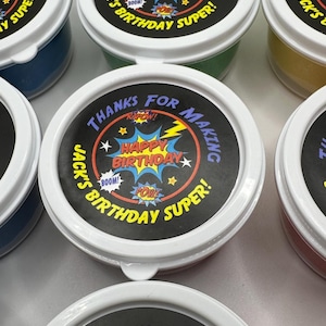 May include: Several small, round containers with white lids. Each lid has a black label with the text "THANKS FOR MAKING" and "HAPPY BIRTHDAY SUPER!" in a comic book style. The containers appear to hold colorful contents.