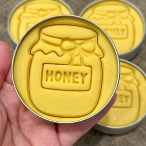 May include: Close-up of four yellow, circular items with a honey jar design. The word "HONEY" is embossed on the jar. The items are in small, silver tins. The background is a neutral color.